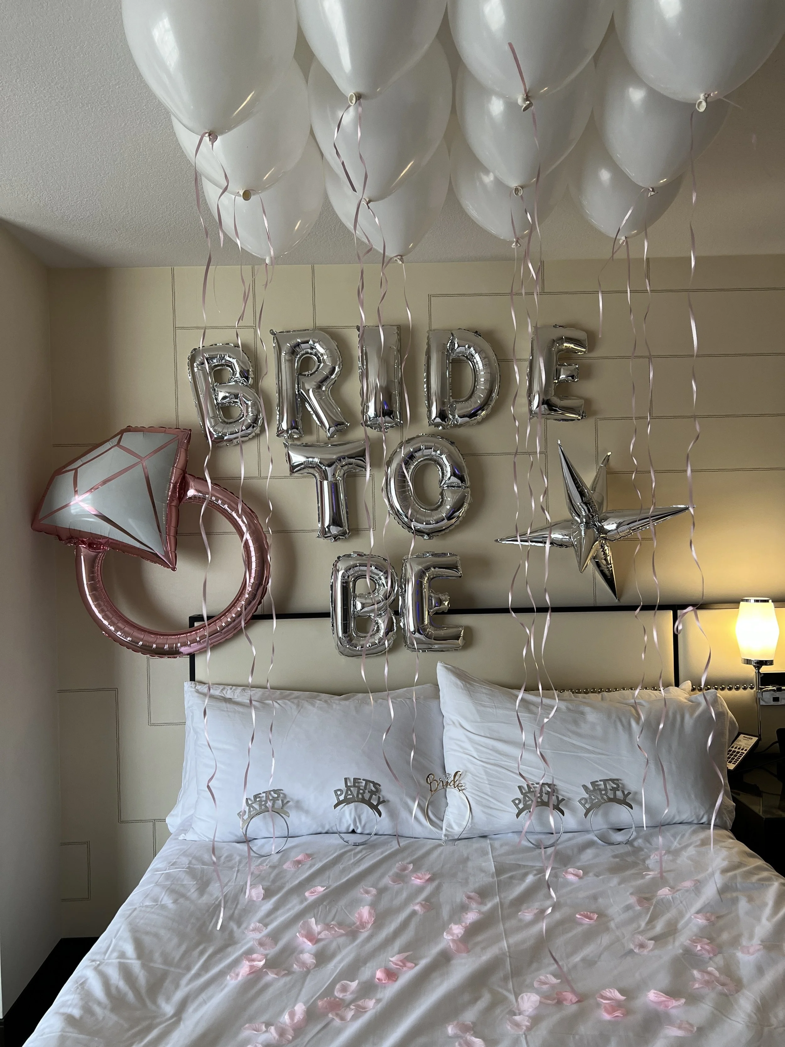 Decorated hotel bedroom with balloons spelling 'BRIDE TO BE', pink rose petals on the bed, and 'Bridal Party' letter decor on pillows.