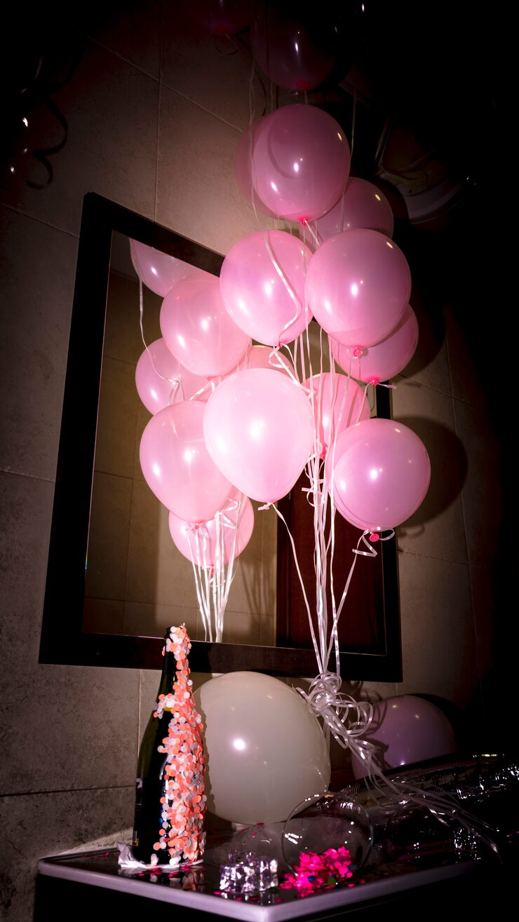 Pink balloons tied together with ribbons, arranged above a table with a decorated bottle, clear balloons, and confetti, displayed in front of a black-framed mirror.