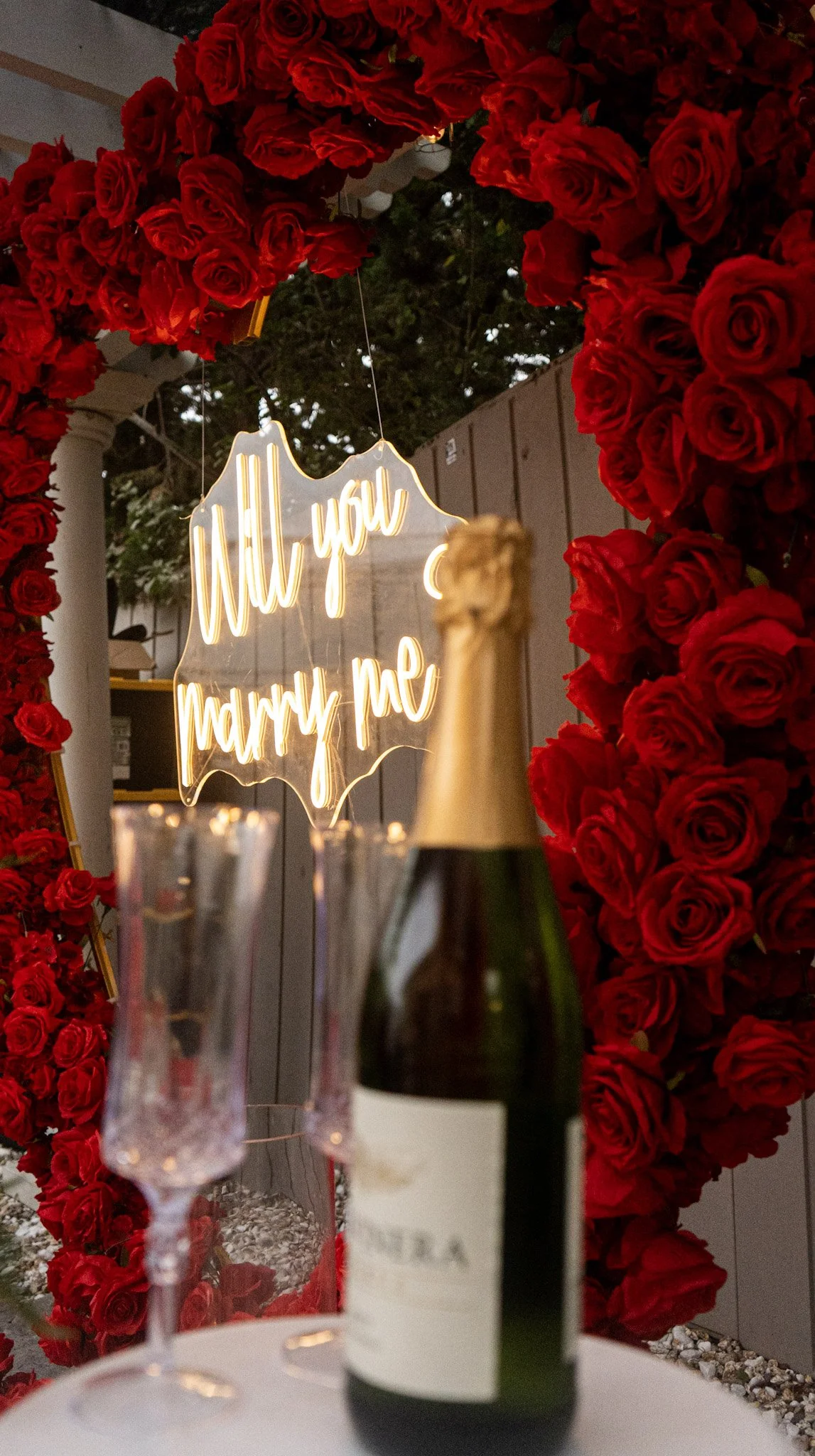 Champagne bottle and two empty champagne flutes on a table in front of a heart-shaped floral arrangement of red roses, with a neon sign that reads 'Will you marry me' hanging above.