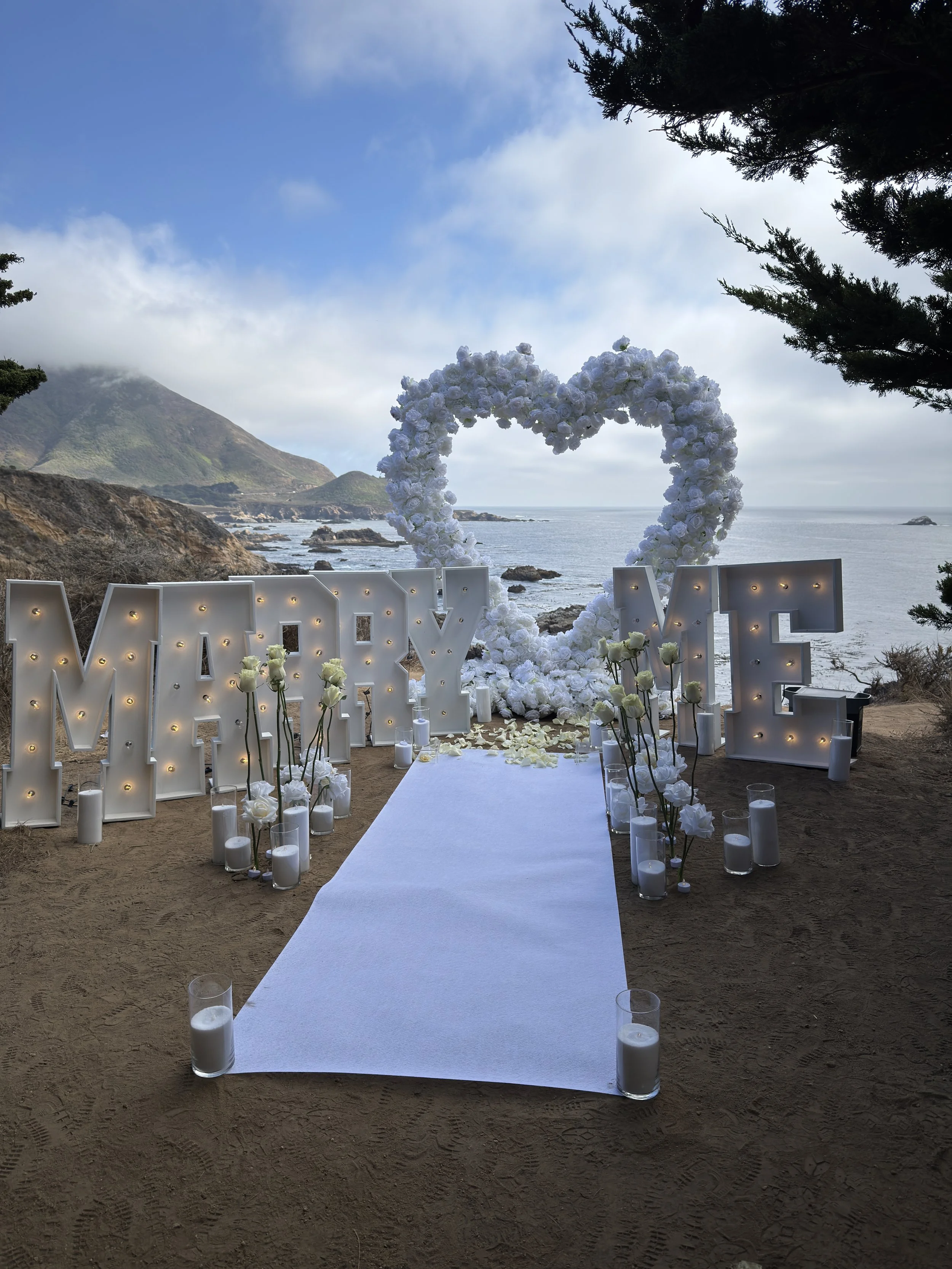 A romantic wedding setup on a beach with white flower arrangements, a heart-shaped floral arch, illuminated 'MARRY ME' letters, candles, and a white aisle runner amid trees with a scenic ocean and mountain background.