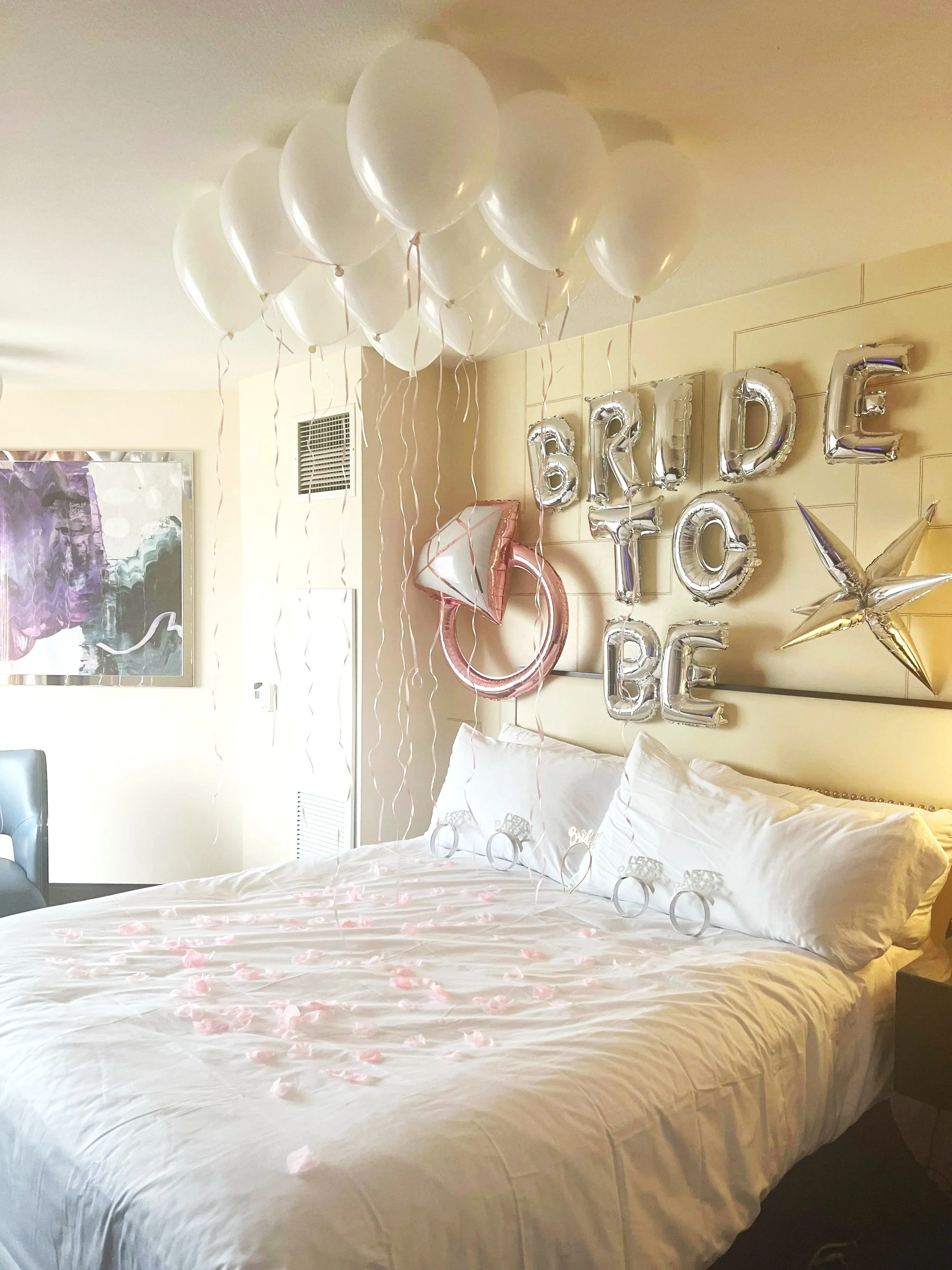 Decorations for a bridal shower with white balloon bouquet, pink and white cake topper balloon, 'BRIDE TO BE' foil balloons on the wall, pink rose petals on the bed, and a star-shaped balloon.