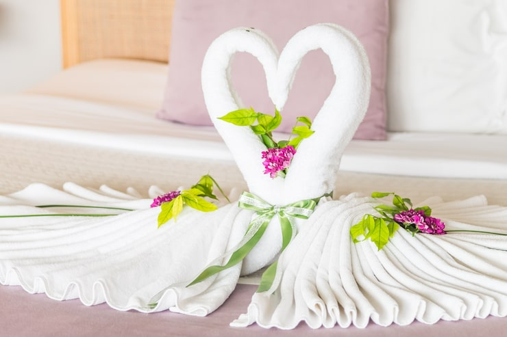Decorative towels folded into swan shapes on a bed with pink and white pillows in the background, adorned with pink flowers and green leaves.
