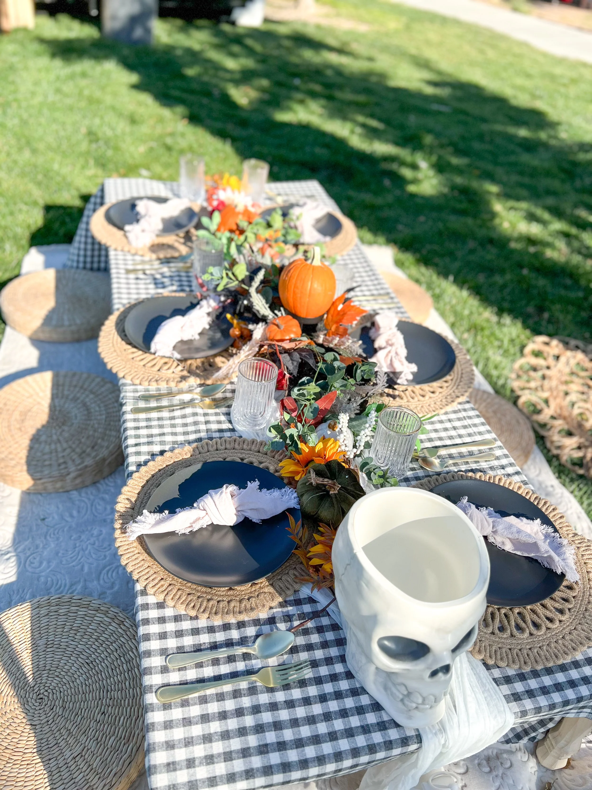 Outdoor dining table decorated with fall-themed decorations including pumpkins, flowers, and faux leaves, set with placemats, black plates, cutlery, and glasses on a checkered tablecloth in a grassy backyard.
