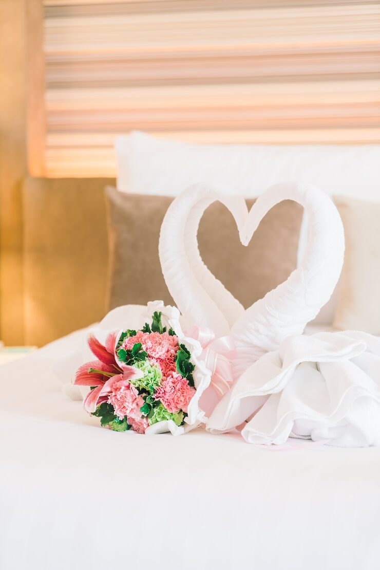 Decorative towel art of two swans forming a heart shape on a bed with a bouquet of pink and green flowers.