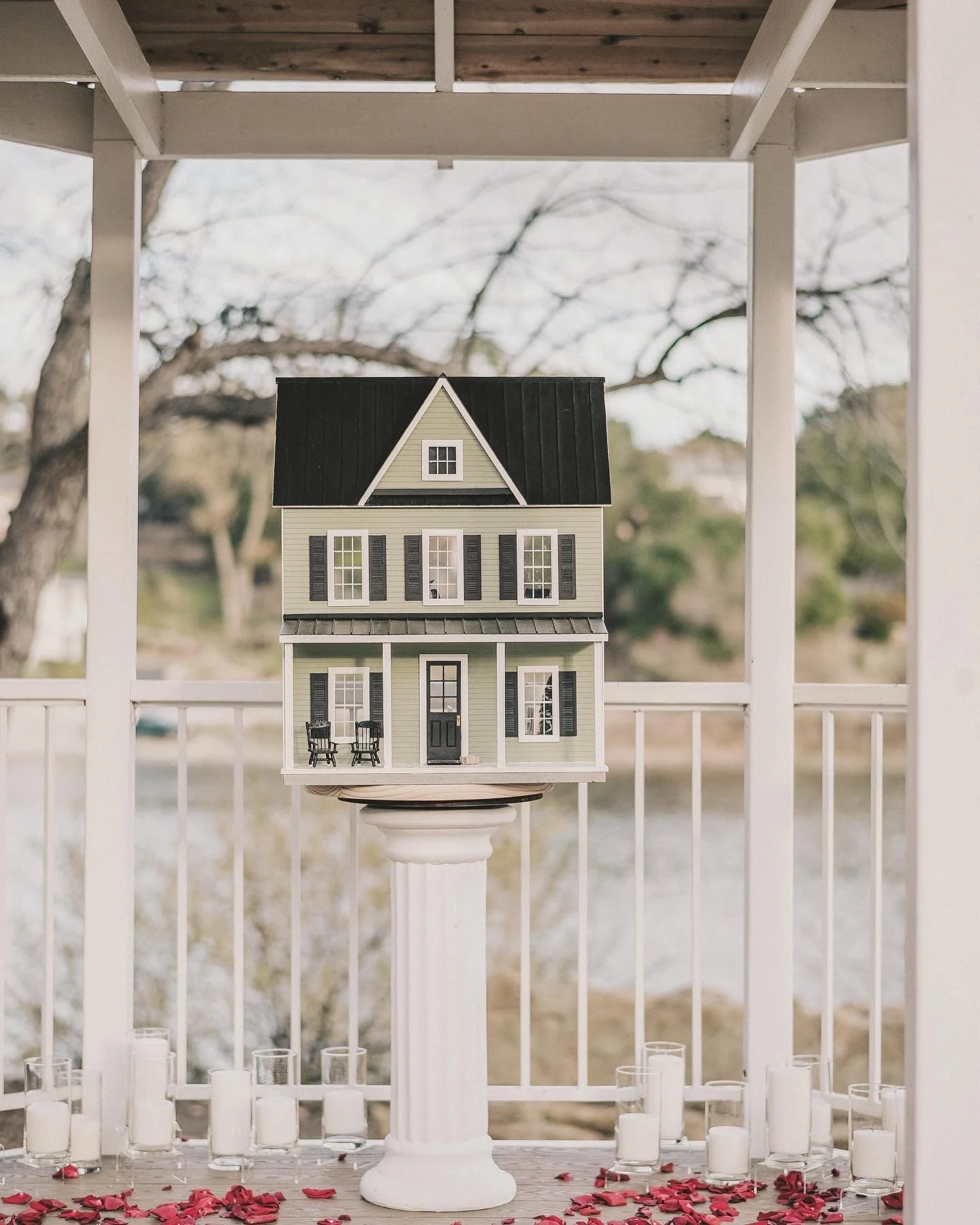 Model of a two-story house with gray and black exterior, black shutters, and a porch with two chairs, displayed on a white pedestal outdoors, with candles and rose petals on the ground.