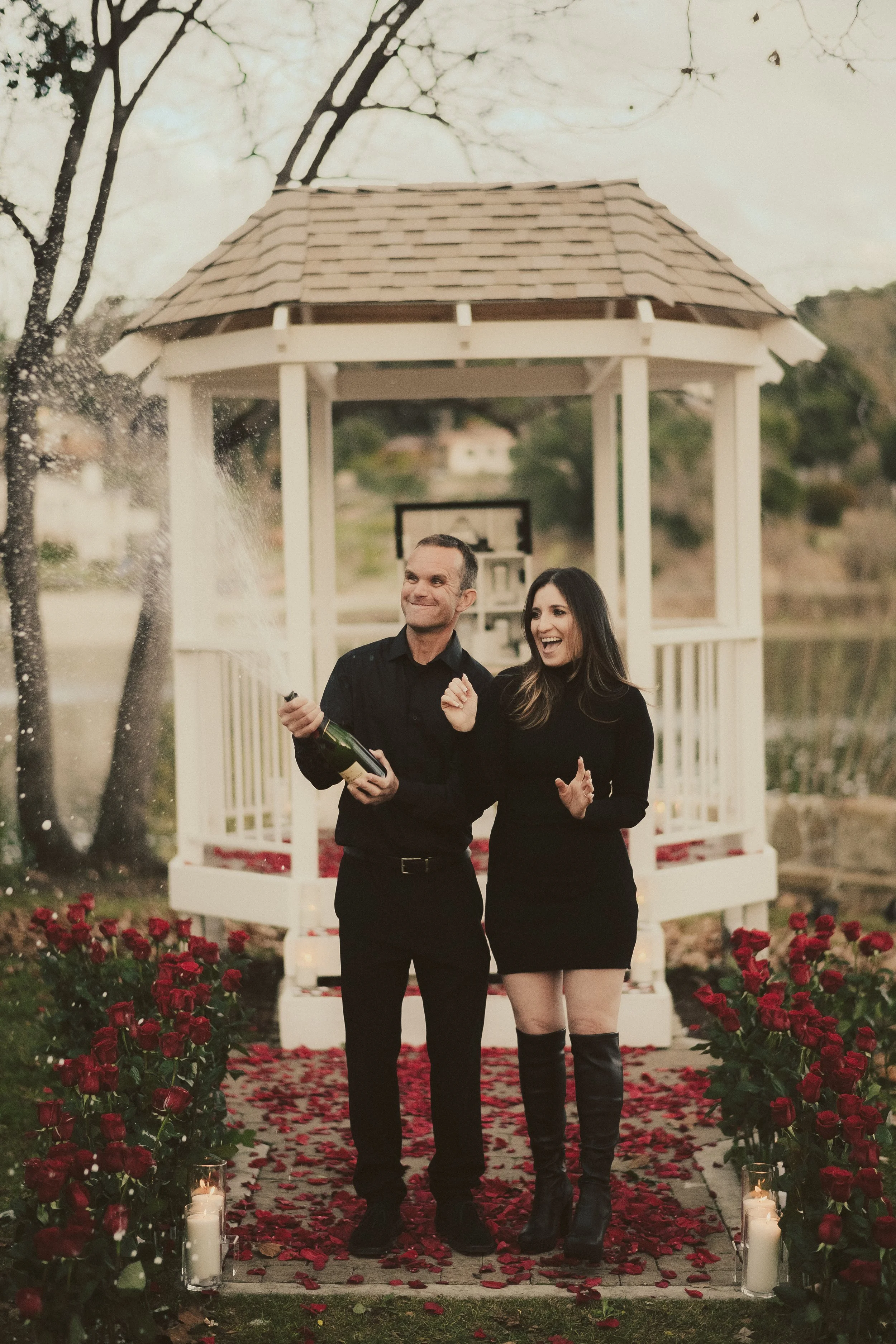 A man and woman celebrating outdoors, standing on a rose petal-lined walkway with candles, near a white gazebo, holding a champagne bottle, with joyful expressions.