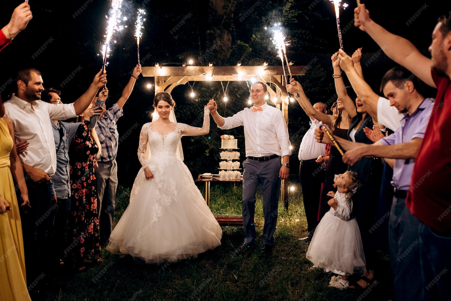 A wedding celebration outdoors at night with the bride and groom holding hands in the center, surrounded by friends and family holding sparklers. There is a multi-tiered wedding cake behind them under a wooden arch decorated with string lights.