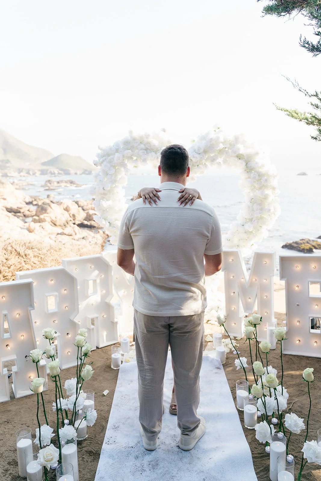 A man and woman embracing during a wedding or romantic ceremony on a beach, with a heart-shaped floral arrangement in the background and the ocean in the distance.