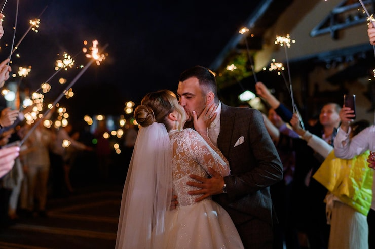 A bride and groom sharing a kiss during their wedding celebration with sparklers held by guests in the background at night.