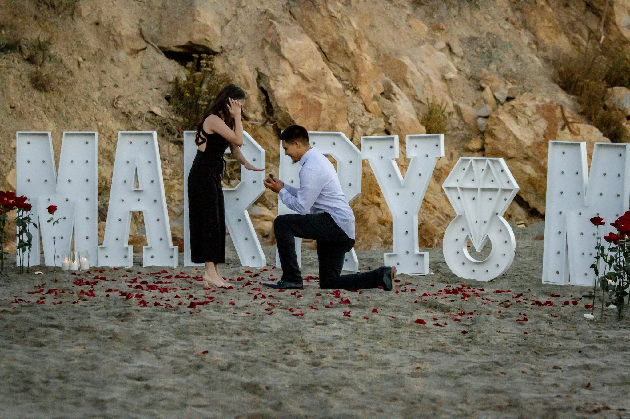 Man proposing marriage to woman on the beach during sunset with large decorative letters spelling 'MARRY' and roses on the ground.