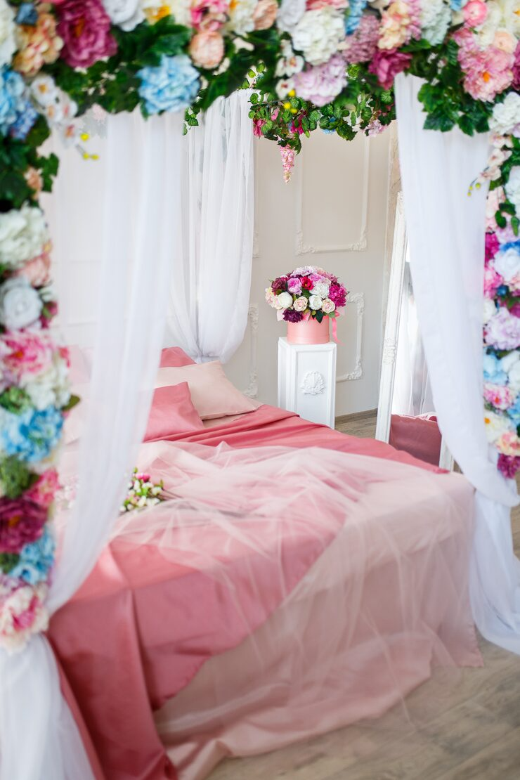 A bed decorated with pink and peach bedding, with floral arrangements and white curtains and drapes surrounding the space, creating a romantic and elegant setting.