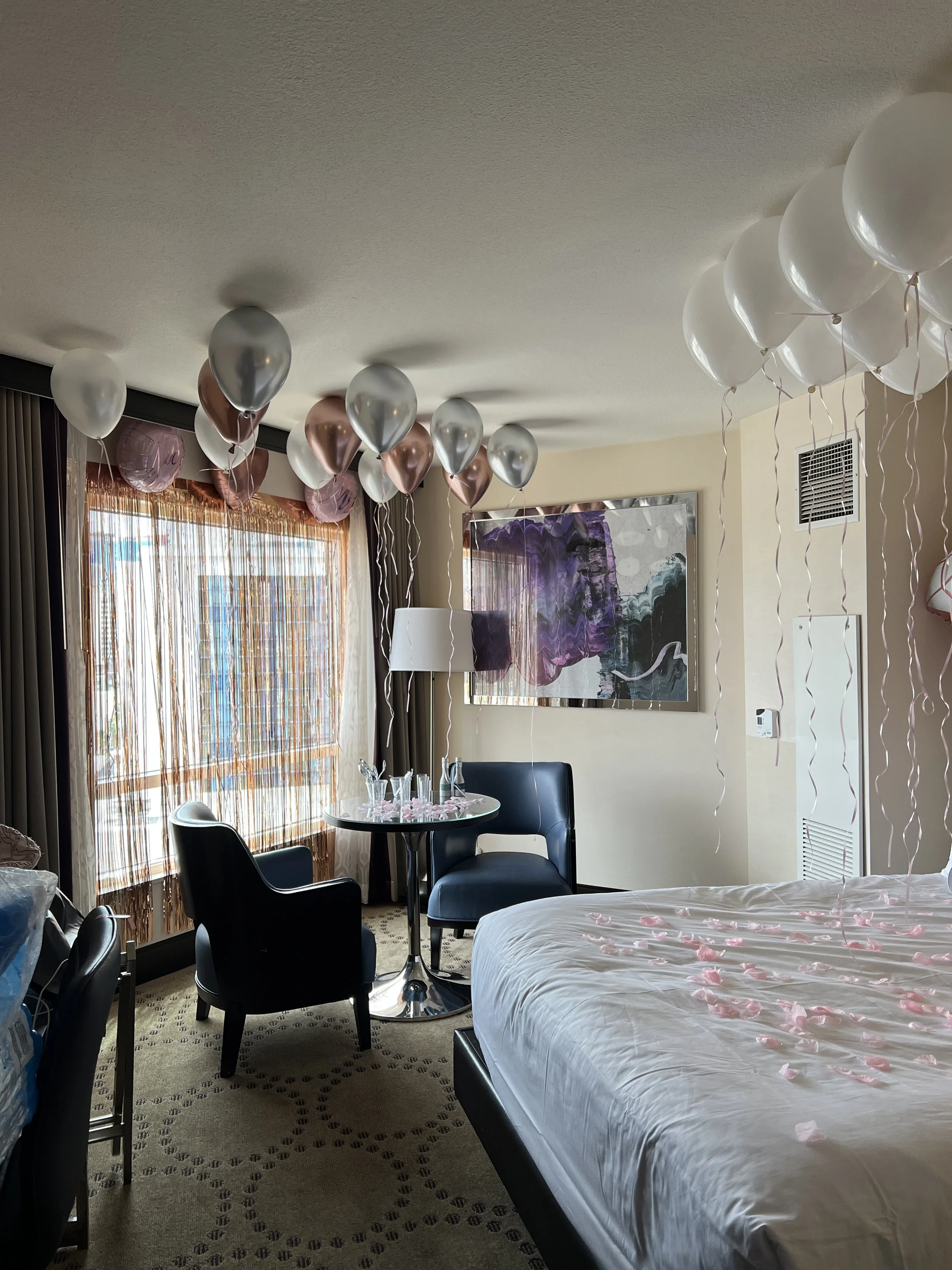 Hotel room decorated with white, pink, and purple balloons, rose petals on the bed, a small round table with glasses, and a painting on the wall.