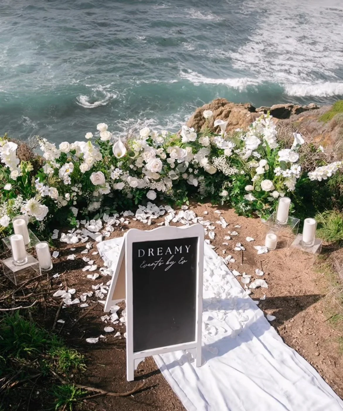 A small wedding altar setup at the edge of a rocky coastline with white flowers and candles, and a black sign that says 'DREAMY Events by E'.