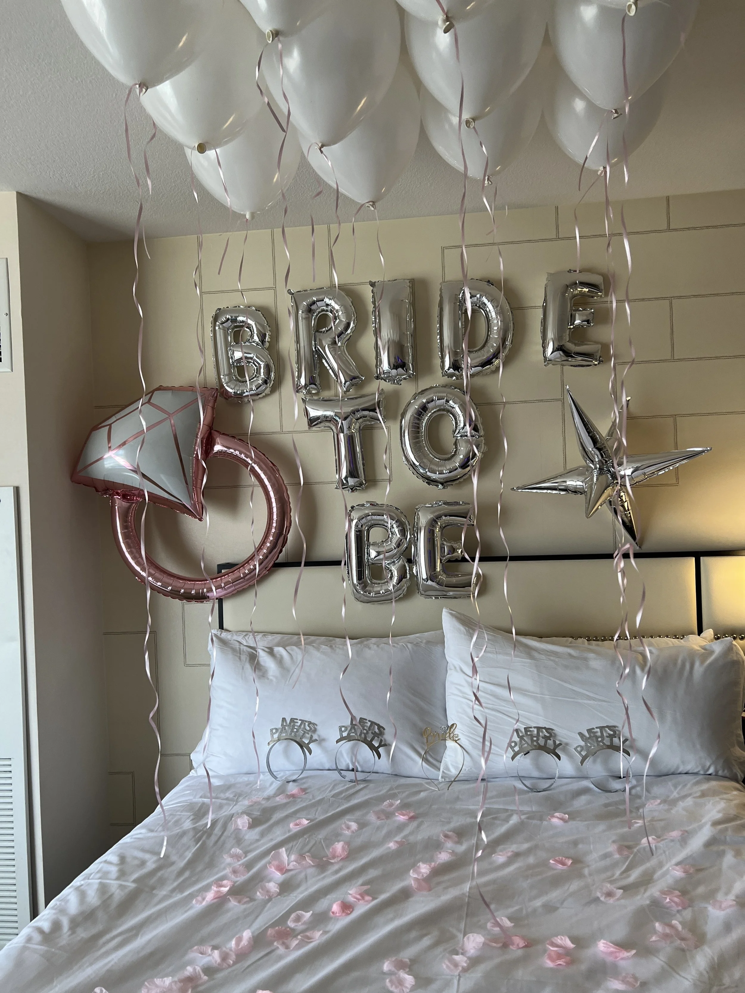 Birthday decorations above a bed, including silver letter balloons spelling 'BRIDE TO BE,' pink and silver star balloons, white balloons hanging from the ceiling, and pink rose petals on the bed.