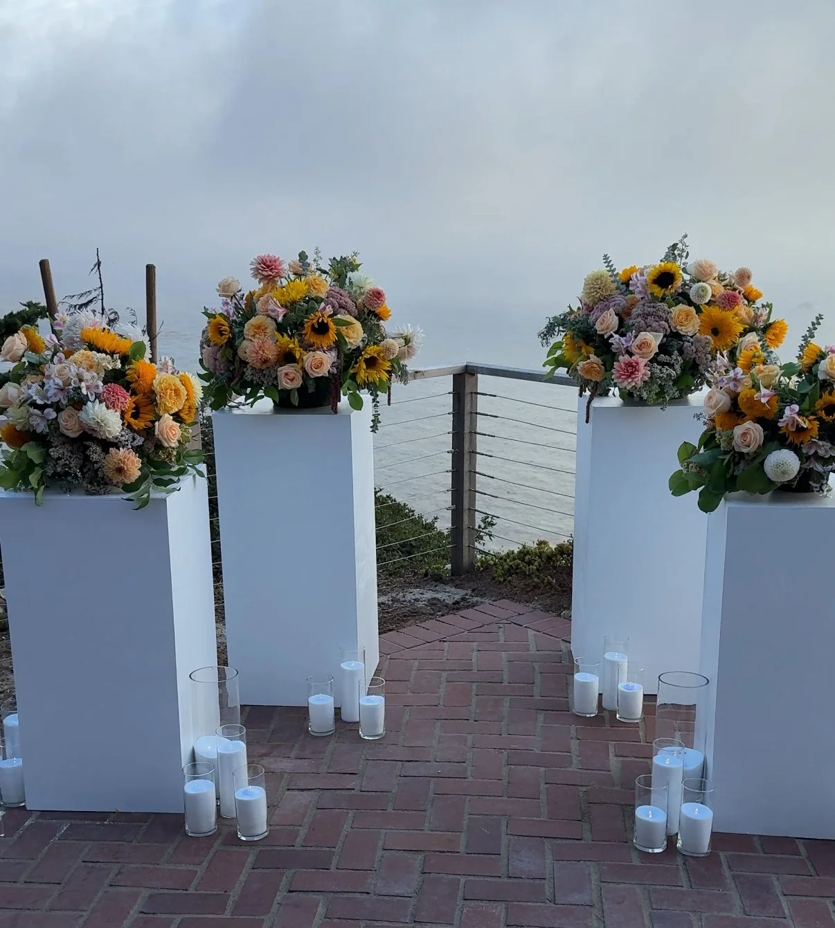 Elegant outdoor floral arrangement setup with flower bouquets on white pedestals, surrounded by candles on a brick pathway, overlooking water.