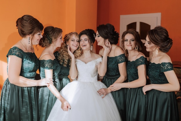 Bride in a white wedding gown with six bridesmaids in dark green dresses inside a house with orange walls.