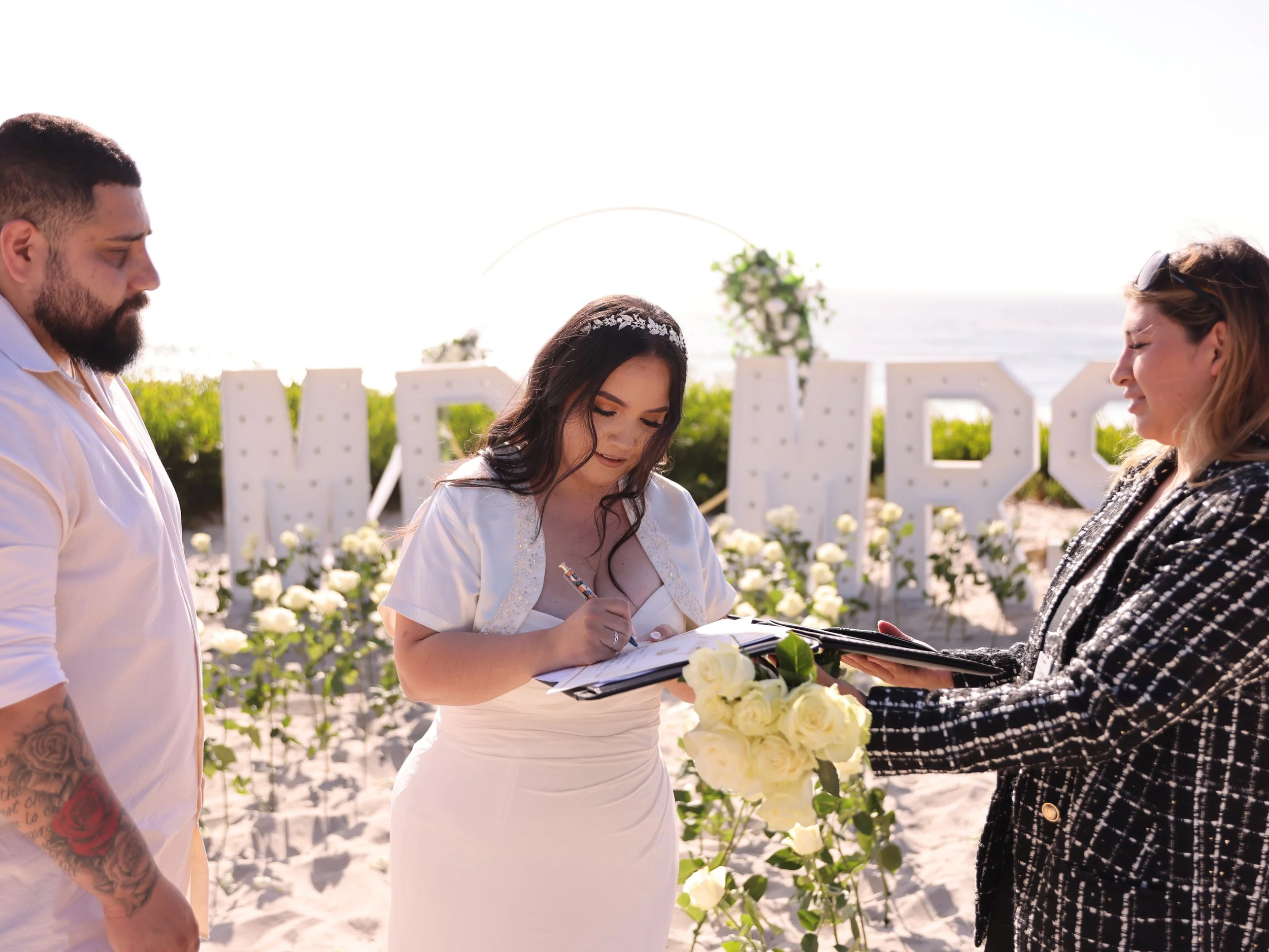 Woman in wedding dress signing marriage license at beach ceremony with man and officiant holding bouquet and nearby decorative letters spelling 'MARC'}
