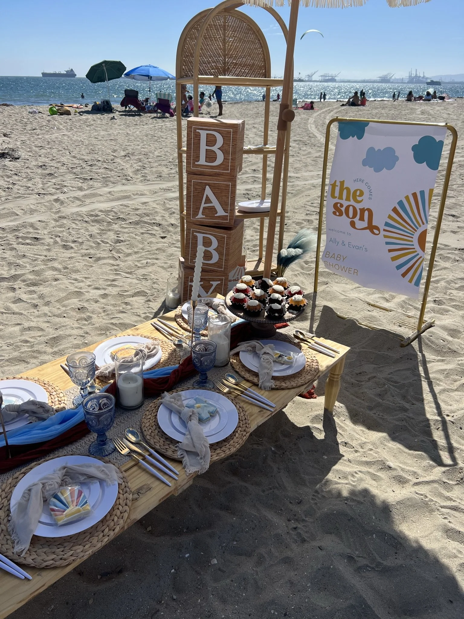 Beach baby shower setup with a wooden table, plates, cups, and cupcakes, near the shore with umbrellas and people in the background, and a sign that says 'the son' for Ally and Evan's baby shower.