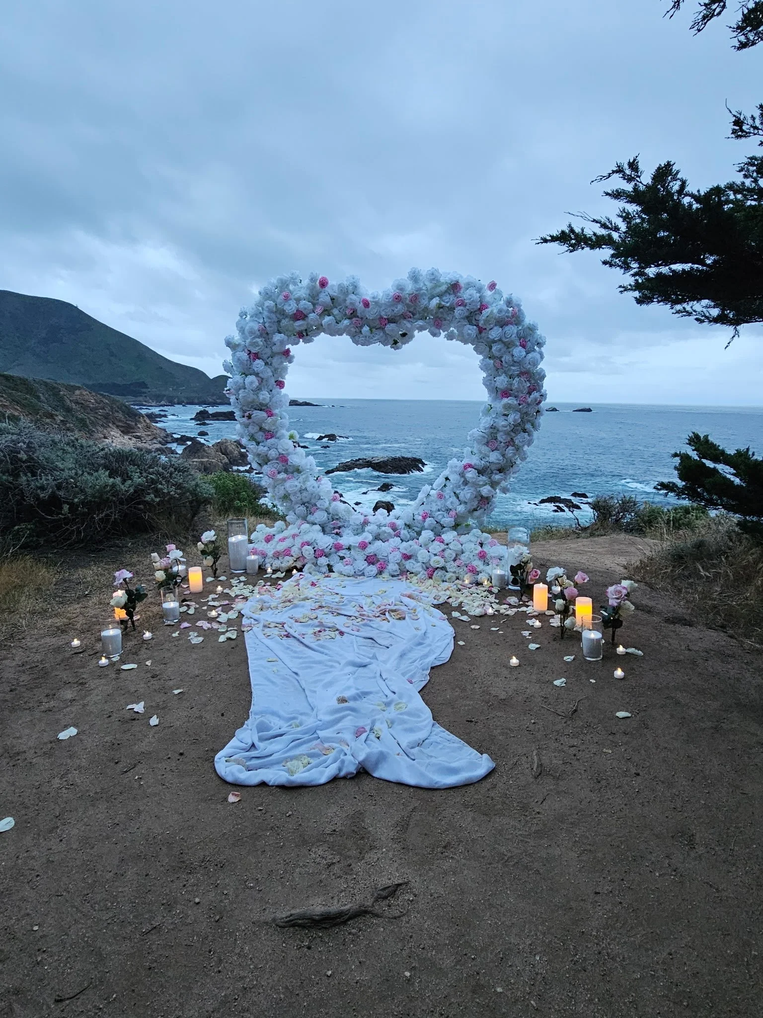 A romantic outdoor wedding setup with a heart-shaped flower arch made of white and pink roses, surrounded by candles and flowers on a dirt path near the ocean with rocky cliffs and cloudy sky in the background.