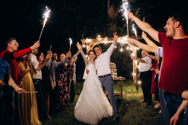 A newlywed couple at their wedding celebration, surrounded by friends and family holding sparklers at night outdoors.