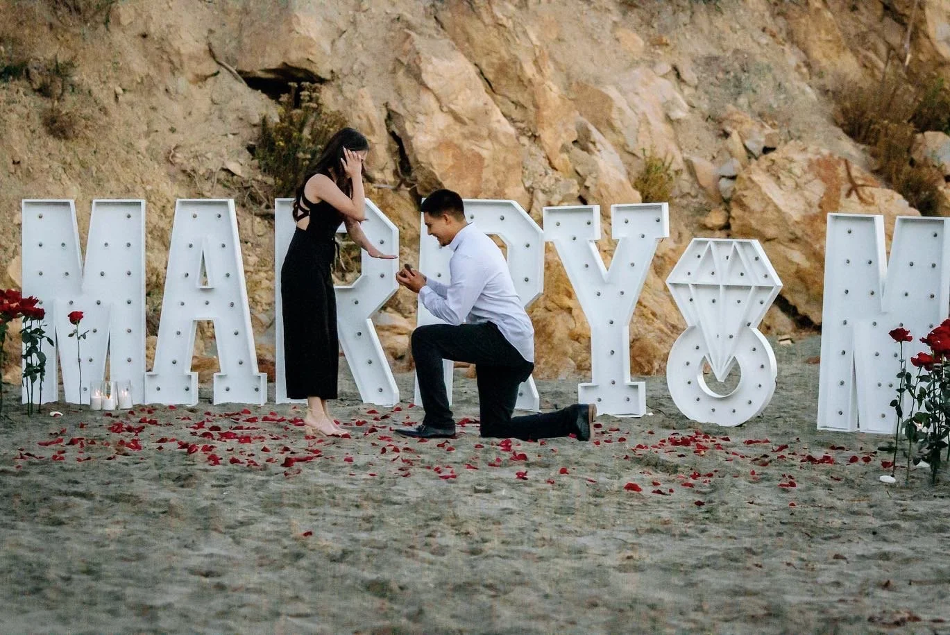 A man proposing marriage to a woman on a beach with large illuminated letter signs spelling 'HAPPY' in the background, scattered rose petals, and candles.