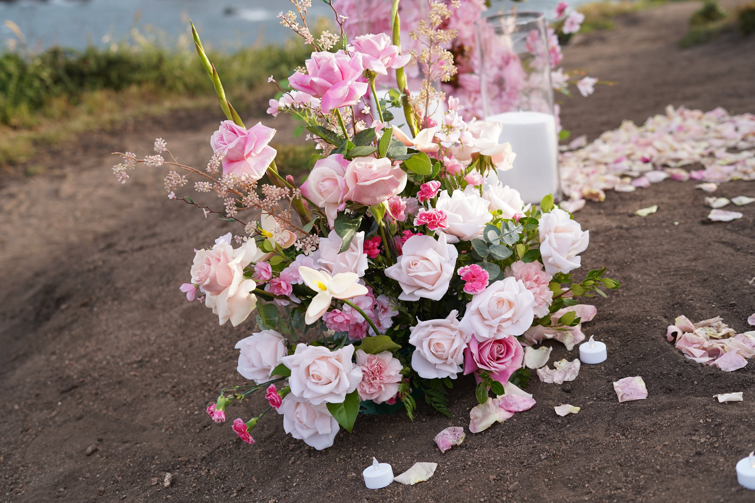 Flower arrangement with pink and white roses, pink carnations, and other flowers on the ground at a memorial site with candles and flower petals.