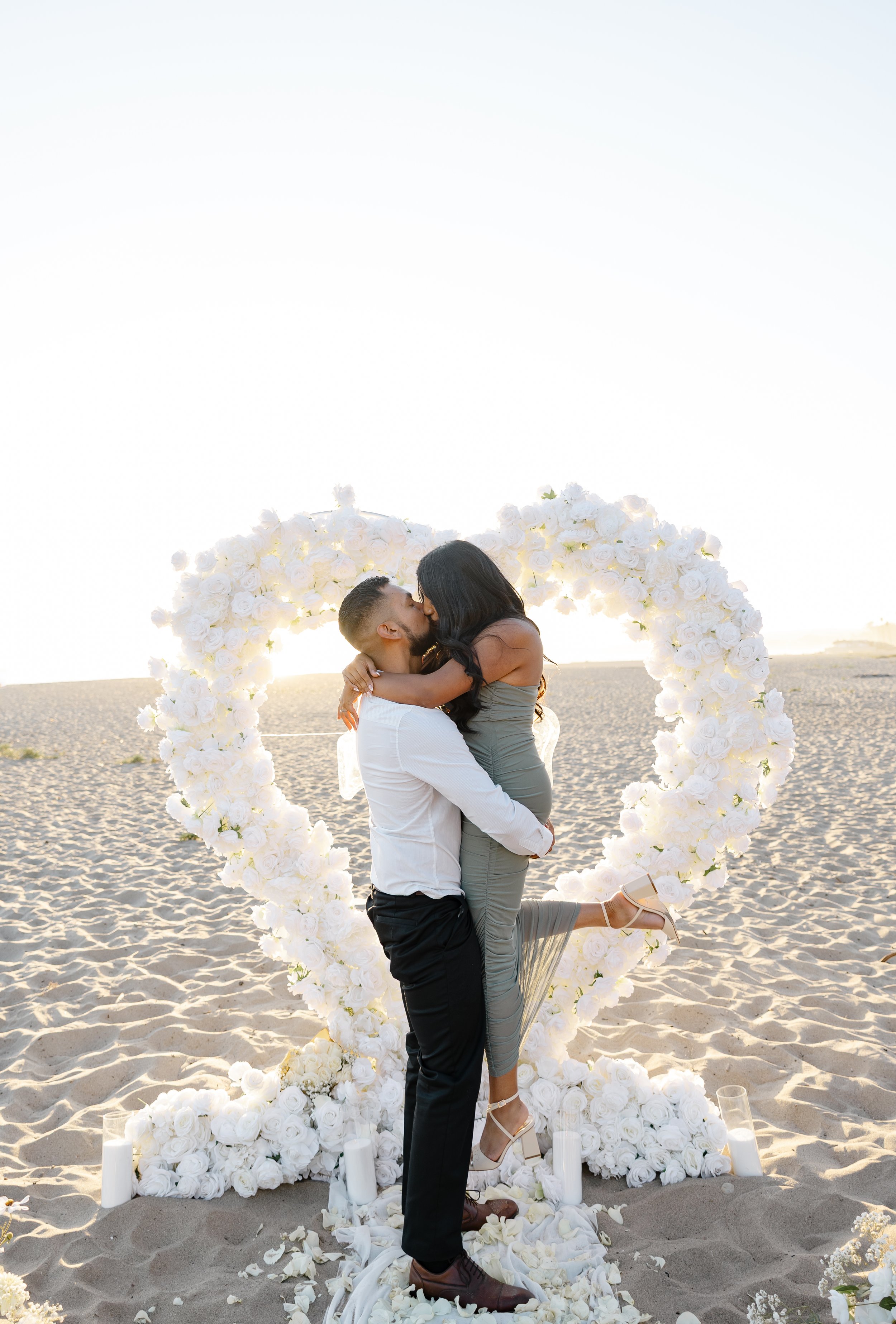 A couple kissing and embracing on a sandy beach in front of a heart-shaped floral decoration during sunset.