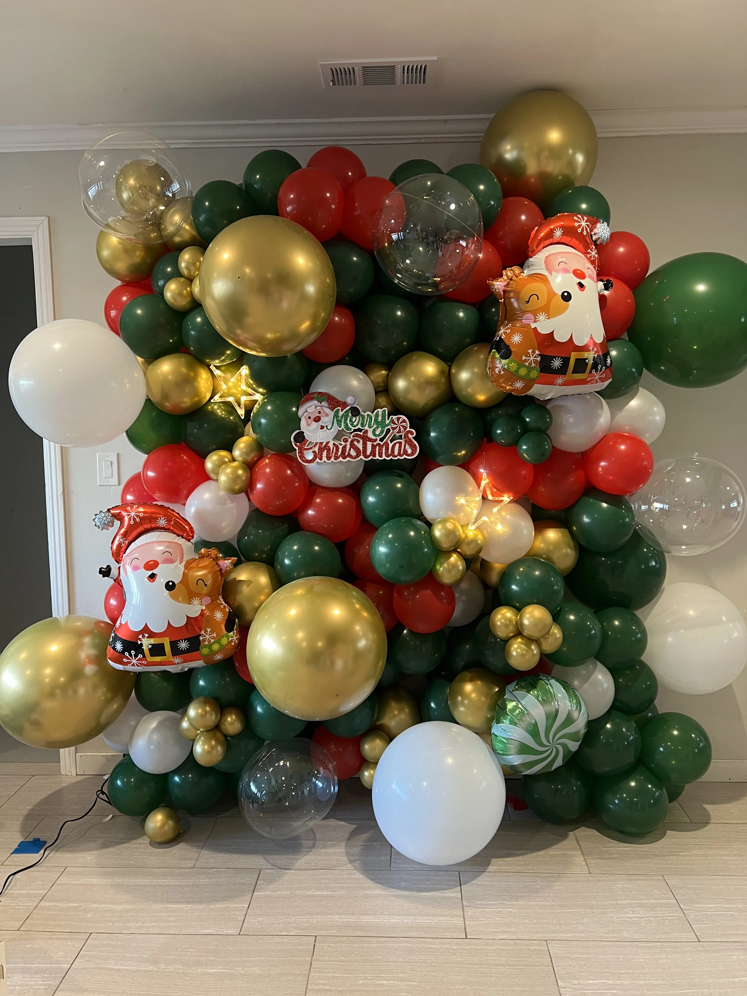Christmas balloon decoration with red, green, white, and gold balloons, including Santa Claus and reindeer foil balloons, a "Merry Christmas" sign, and string lights.