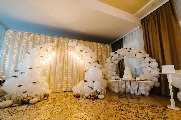 White balloon arch and decorations with a tiered cake on a table, decorated for a celebration or event, in a room with curtains and string lights.