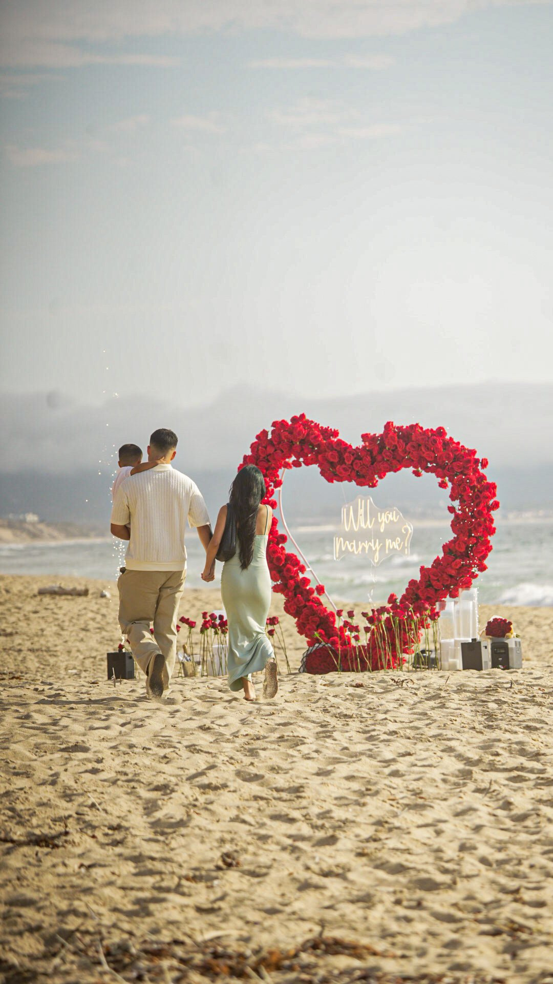 A couple holding hands and walking on a beach towards a heart-shaped floral decoration with a neon sign that says 'Will you marry me?' at sunset.