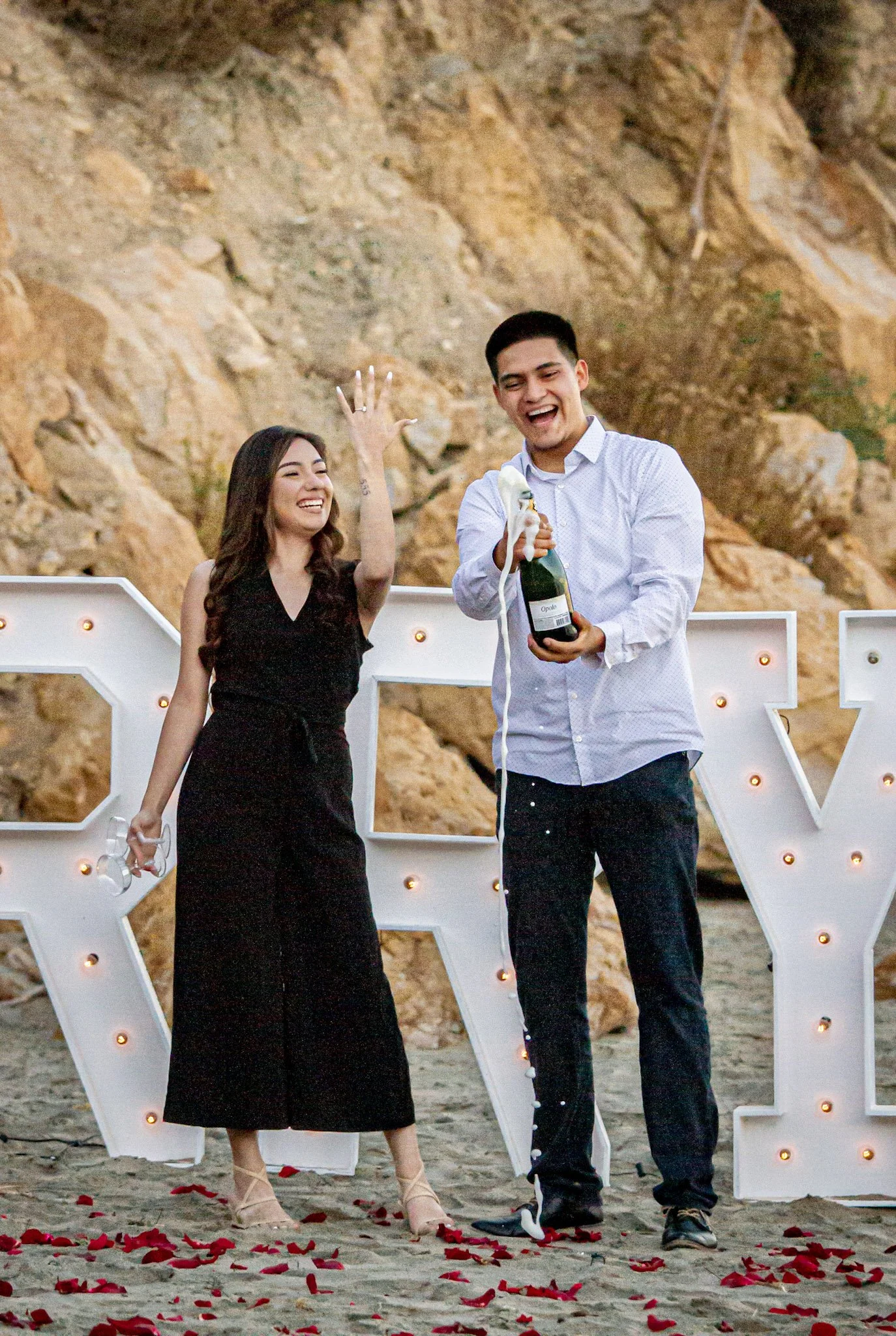 A couple celebrating on a beach, with a woman in a black dress and a man in a white shirt, opening a champagne bottle, surrounded by New Year decorations and rose petals.