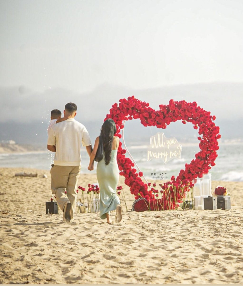 A couple holding hands and walking on the beach at sunset, with a child in their arms, towards a large heart-shaped floral arrangement with a sign that says "Will you marry me?"
