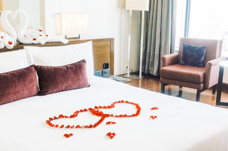A hotel room with a bed decorated with rose petals arranged in the shape of two overlapping hearts. The room has a wooden headboard, two maroon pillows, swan-shaped towels on the bed, a pink armchair with a dark pillow, a side table, and a large window with curtains.