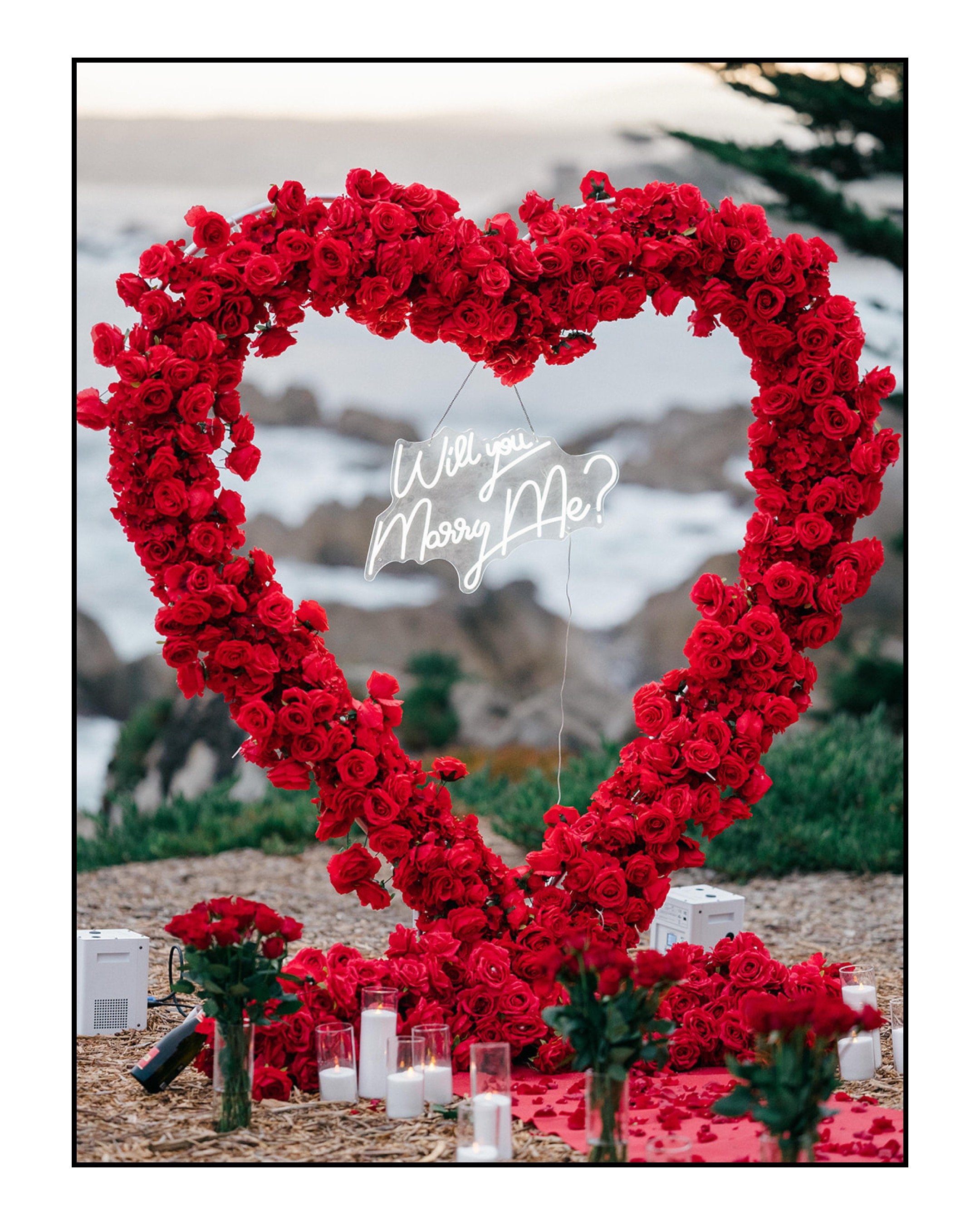 A romantic outdoor proposal setup with a heart-shaped frame covered in red roses, a neon sign reading 'Will you marry me?,' candles, and flower arrangements.