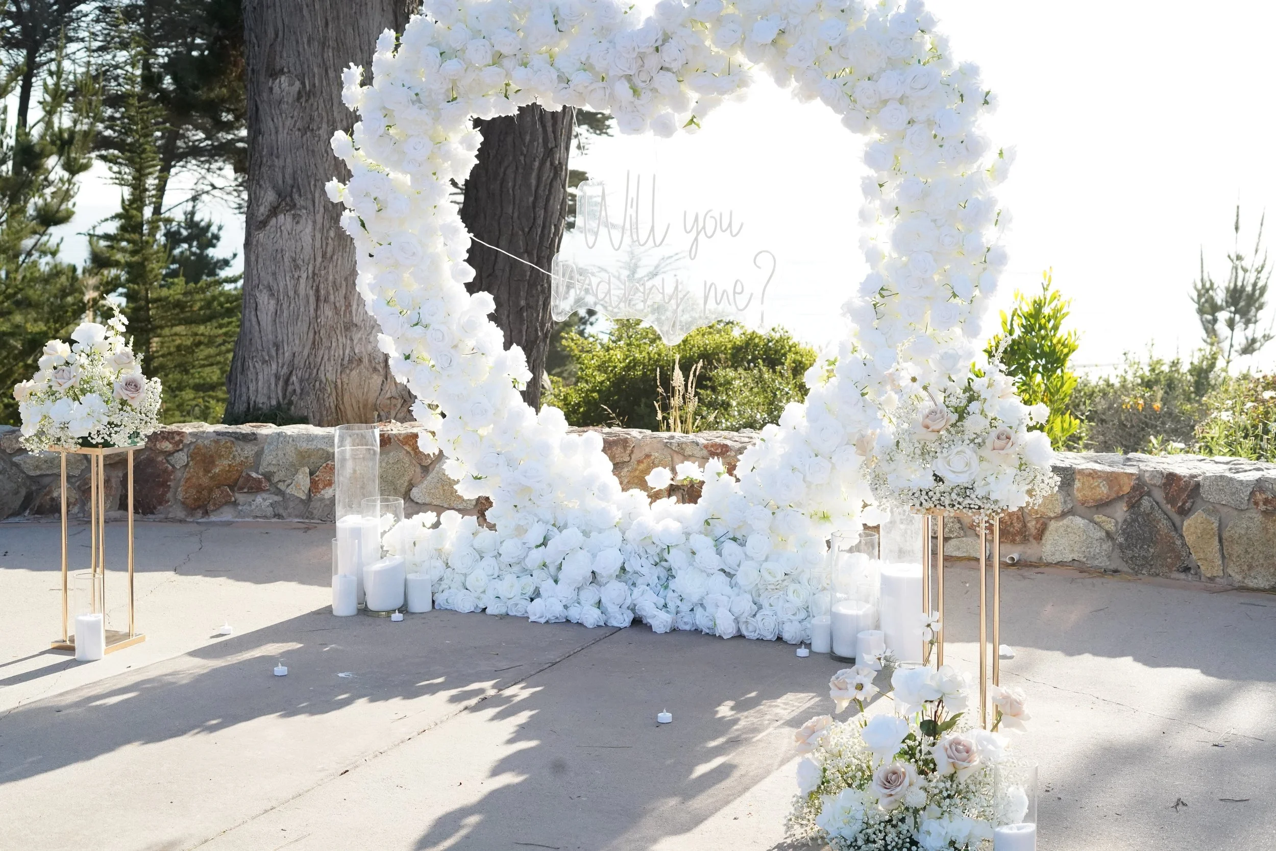 A white floral arch with roses and candles in an outdoor setting, with trees and a stone wall in the background, and a transparent sign that says 'Will you marry me?' hanging in the center.