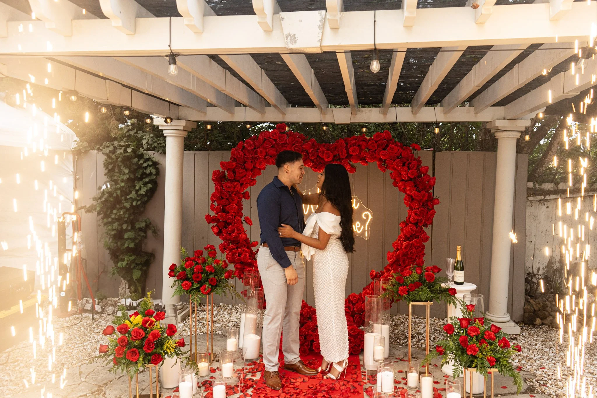 A couple standing close together inside a heart-shaped frame made of red roses, in an outdoor Valentine's Day or romantic celebration setting with candles, flowers, and sparklers.