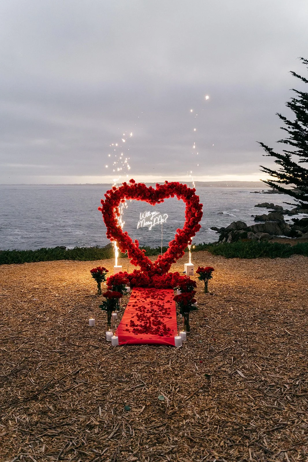 A romantic setup near the ocean featuring a large red heart-shaped floral arrangement and a red carpet with rose petals, surrounded by candles and small bouquets, against a cloudy sky and rocky shoreline.
