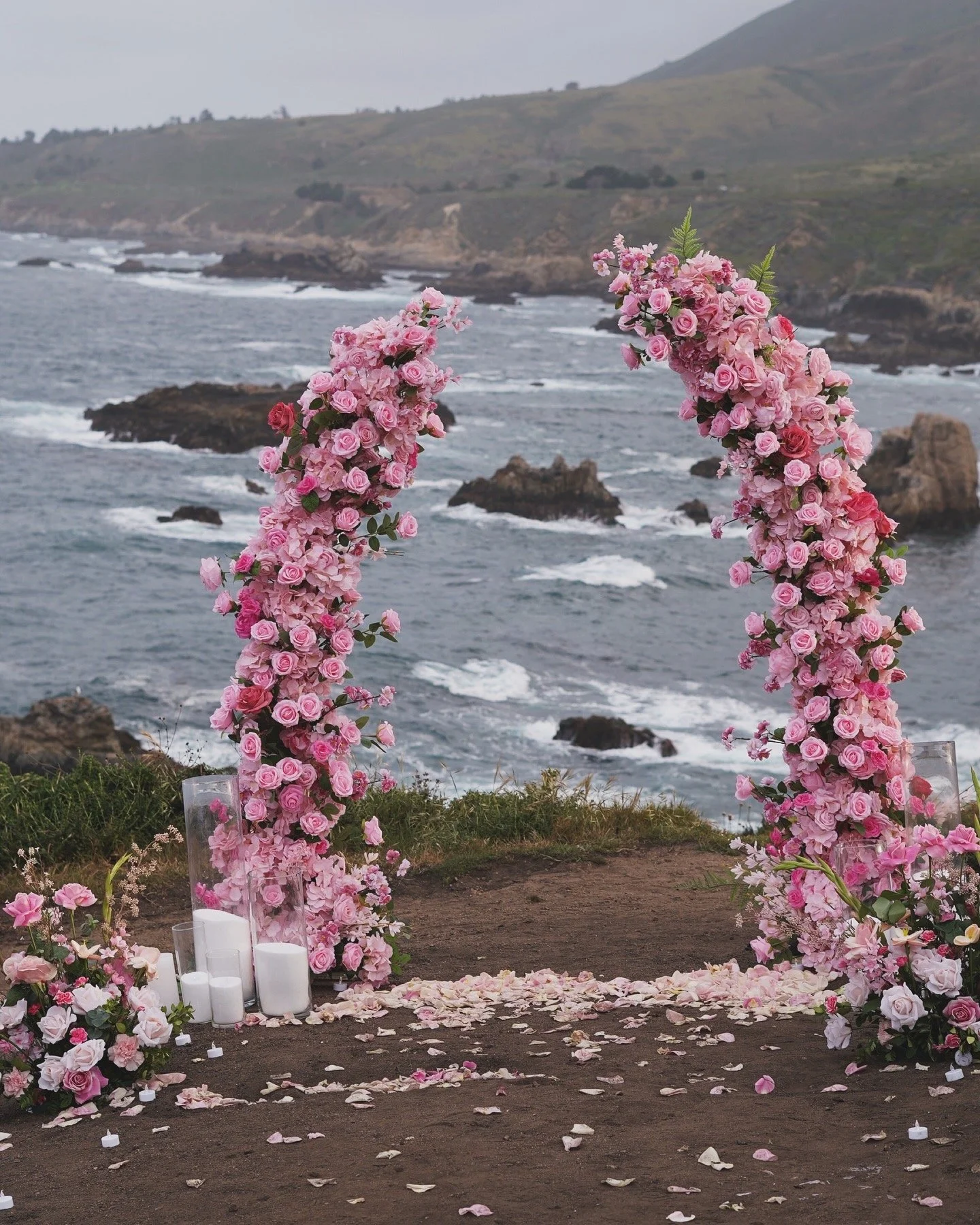 A wedding arch decorated with pink roses and flowers overlooking the ocean at a coastal location.