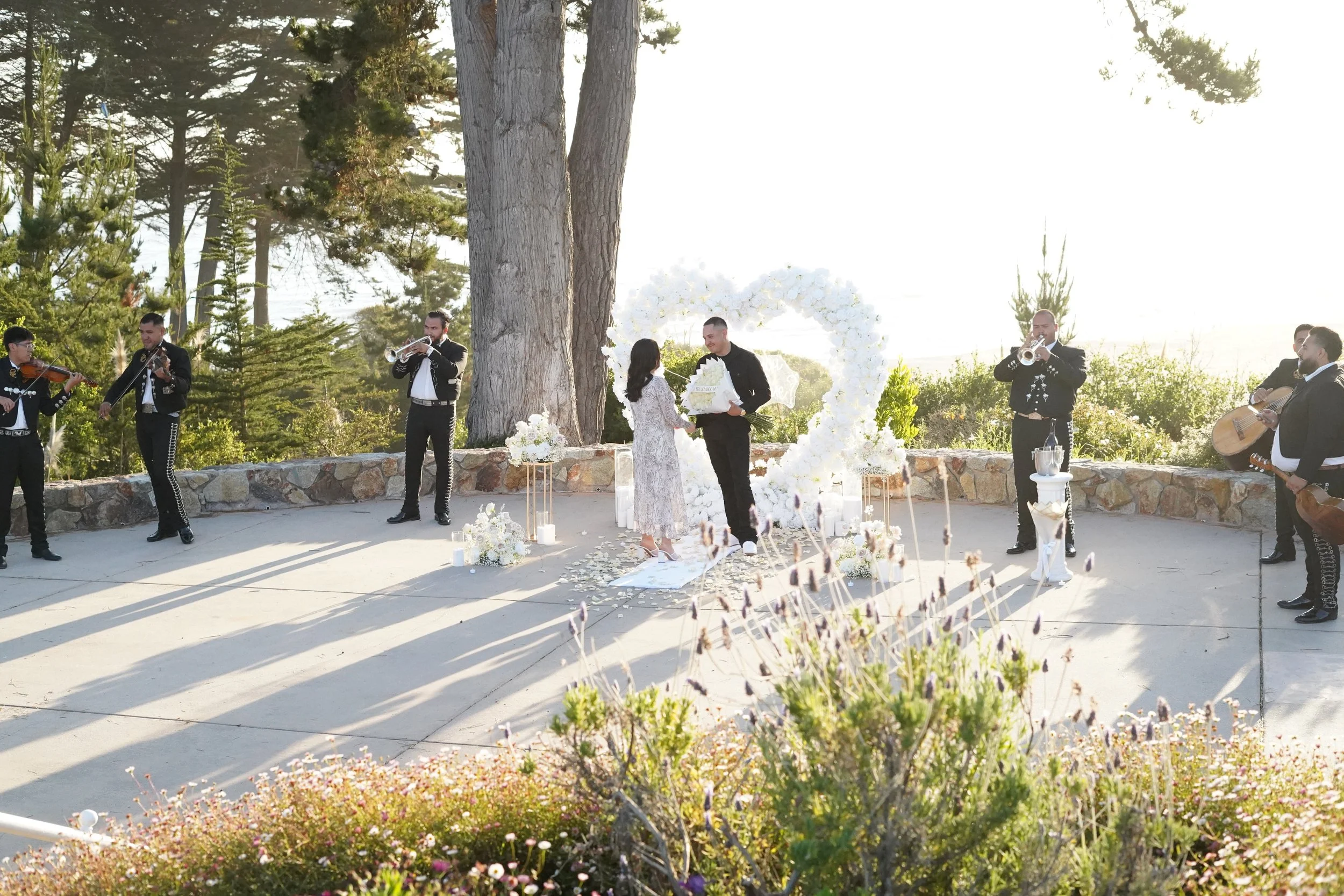 A wedding ceremony outdoors with a couple exchanging vows in front of a heart-shaped floral arch. Musicians are playing instruments nearby, with sunlight and trees in the background.