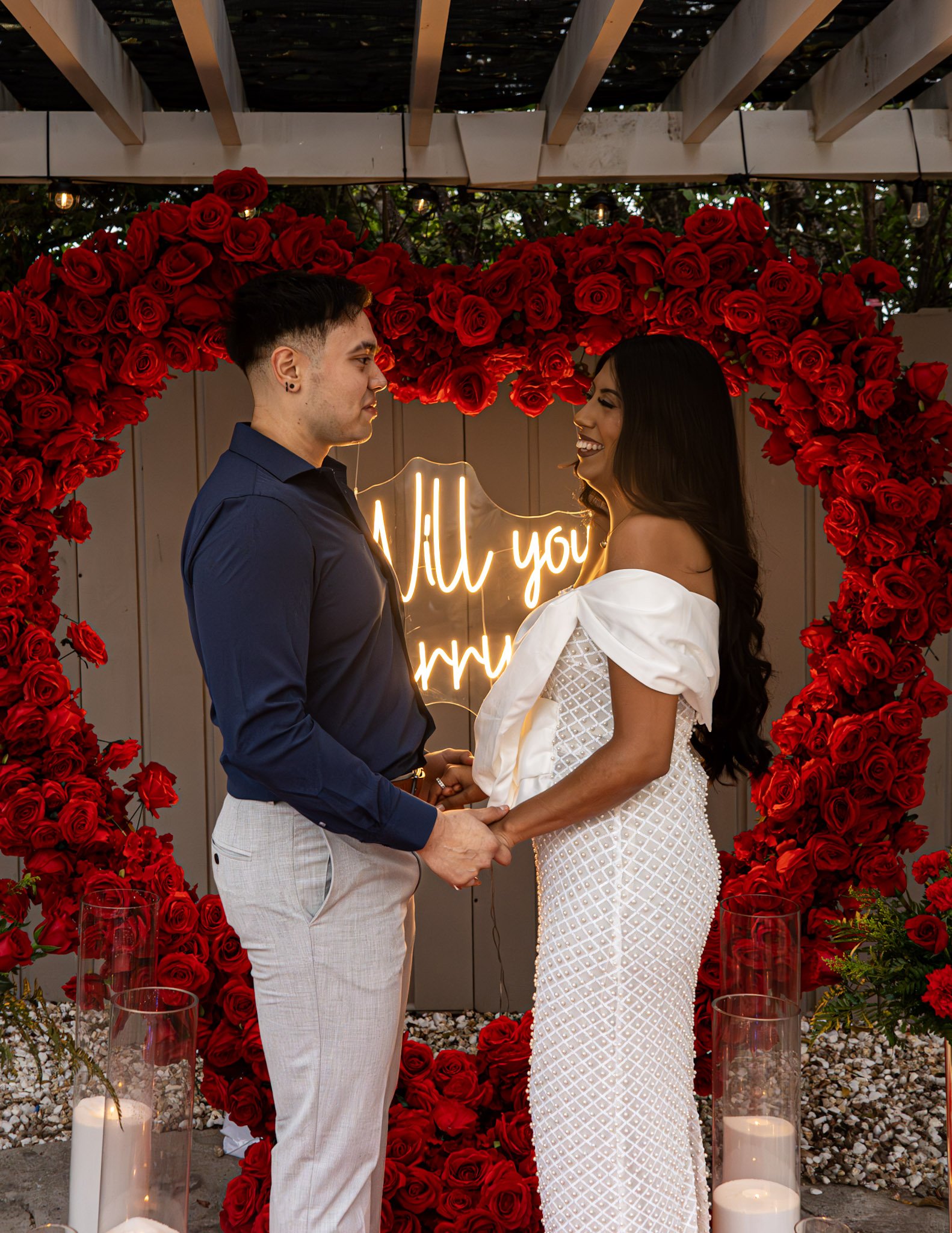 A couple holding hands and looking at each other, standing in front of a heart-shaped rose wall with a neon sign that reads 'All you'.