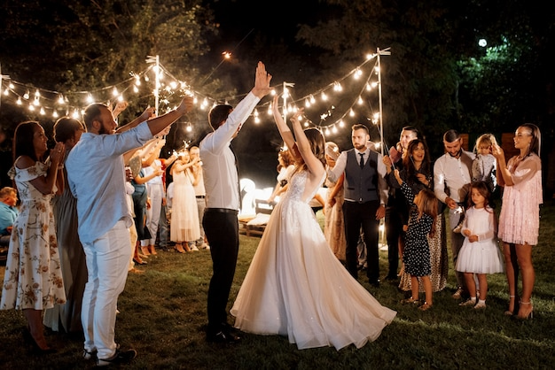 Couple's wedding dance outdoors at night under string lights, surrounded by family and friends, with children and guests watching.