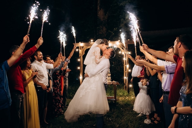 Bride and groom sharing a kiss under an archway at night, surrounded by friends and family holding sparklers.