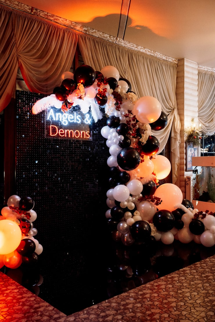 Decorative balloon arch with black, white, and beige balloons and red floral accents in front of a black sequin backdrop with neon sign reading 'Angels & Demons' at an indoor event.