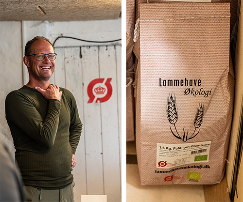 Mand smiler og står ved siden af en hvid væg med et rødt logo bag sig, og der er en pose med lammekød til økologi på hylde til højre.