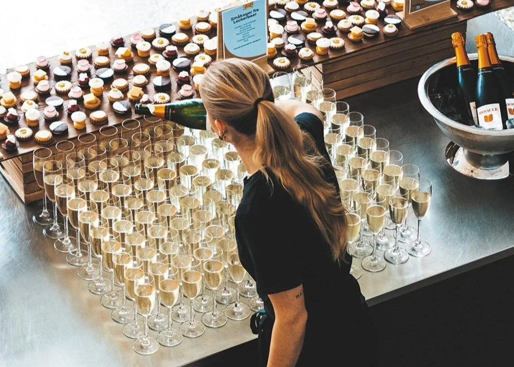 En kvinde lader champagne flyde i glas foran en stor skærm med små kager online.