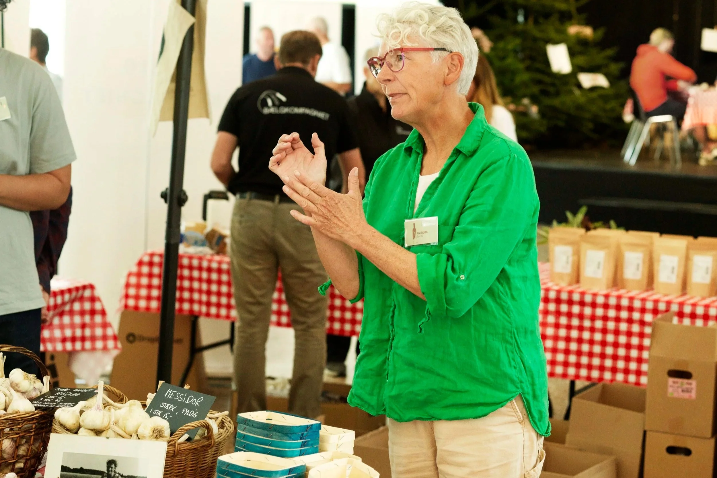 Farmers Market hos Gastronomiet. Hvidløgssælger står ved en bod med produkter,  med flere mennesker i baggrunden.