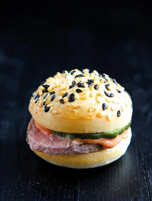 En burger med lyst sesambolle, bøf, syltede agurker og dressing på sort baggrund.