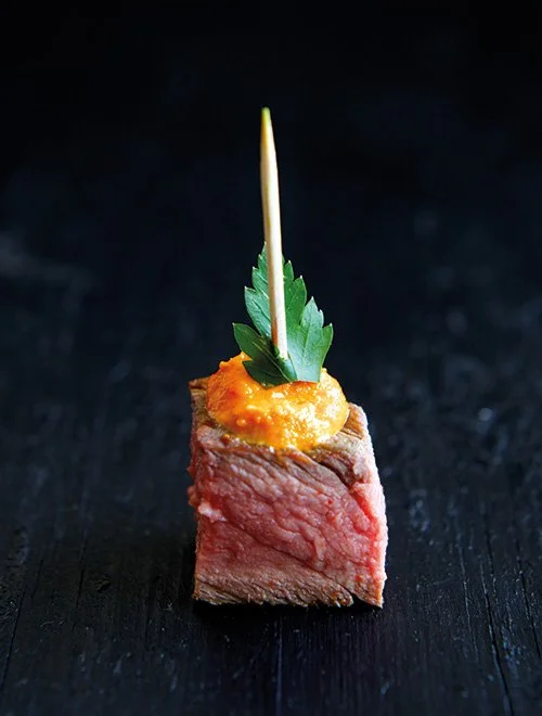 En lille servering af oksekød med en orange sauce og et persilleblad, spiddet med en tandstikker, på en mørk baggrund.