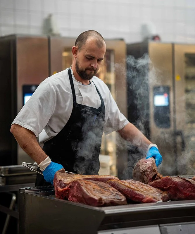 Firmafrokost fra Gastronomiet. Kok står ved en grill med store stykker kød, som han tilbereder, iført kokketøj.