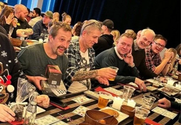 En gruppe mennesker sidder ved et bord og nyder en øl, mens de spiller spil eller deltager i en social aktivitet i en indendørs setting.